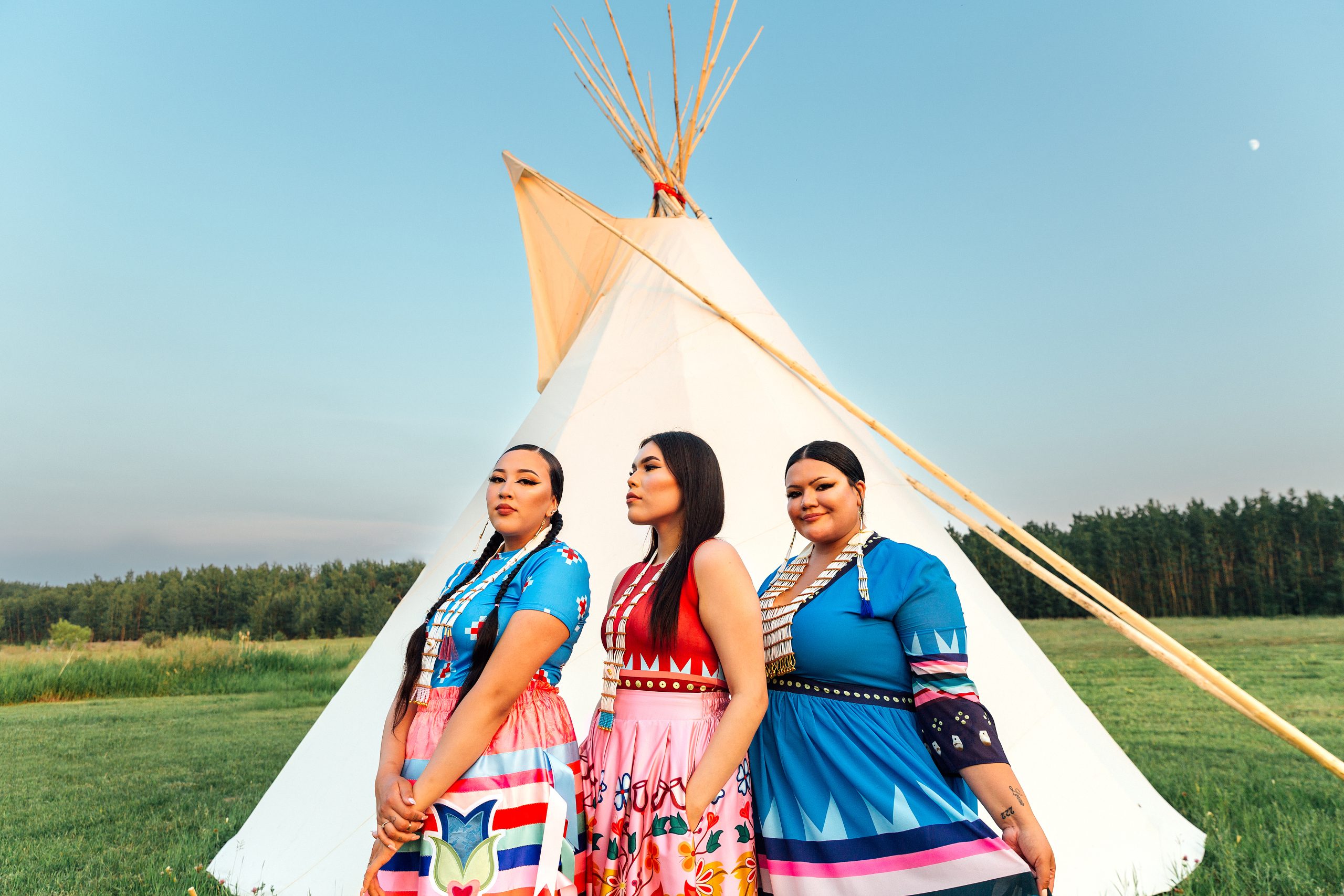 three woman in  Maskawitehew designs standing on green grass with a tipi and trees behind them