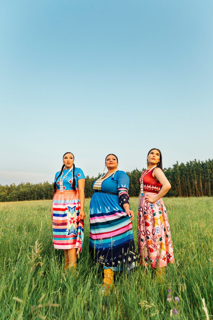 three woman in  Maskawitehew designs standing on green grass with a tree behind them