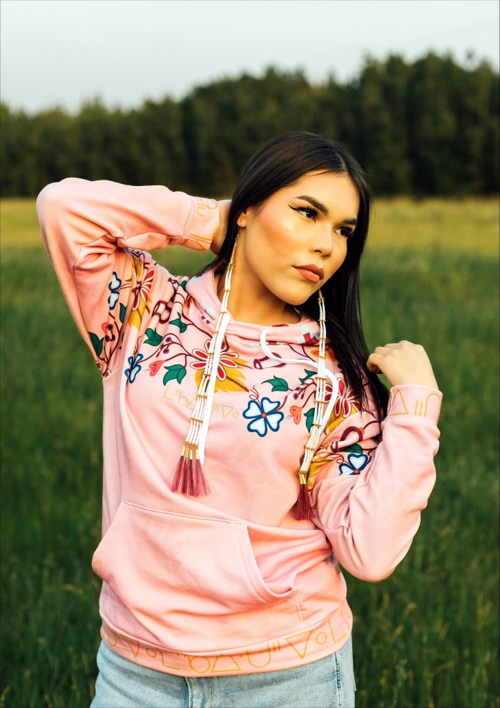 woman in Maskawitehew design cheyenne pink hoodie standing on green grass with trees behind her