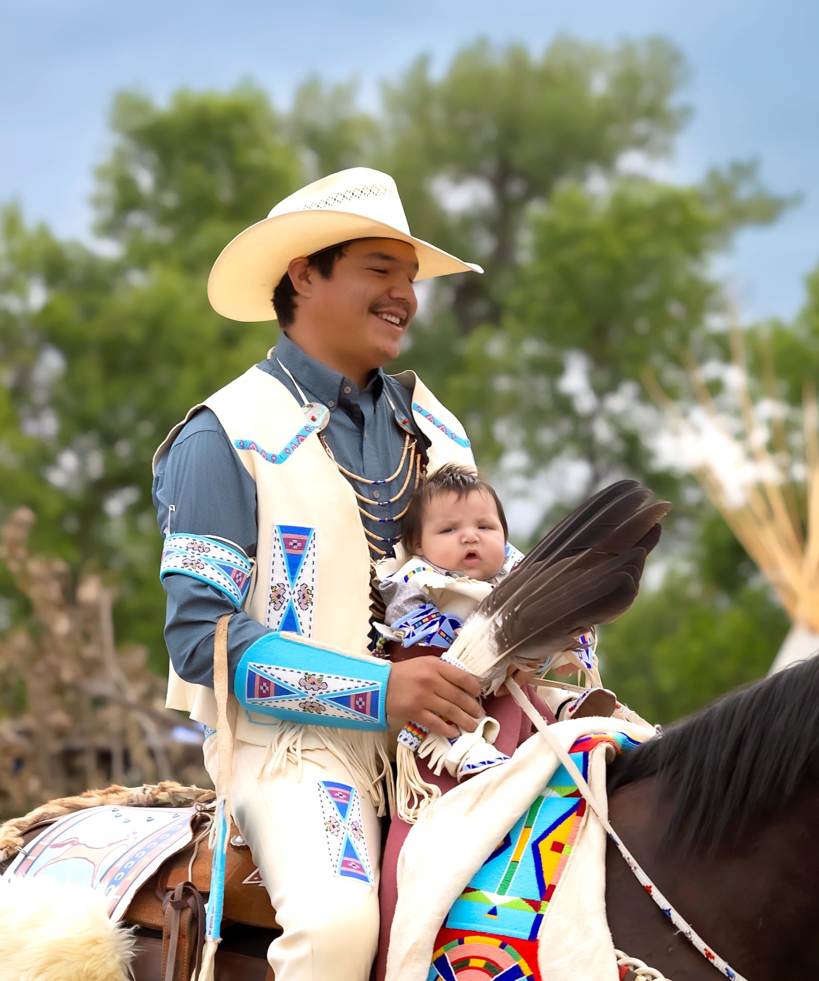 Man in a cowboy hat and beaded vest holding an infant on horseback at Crow Fair.