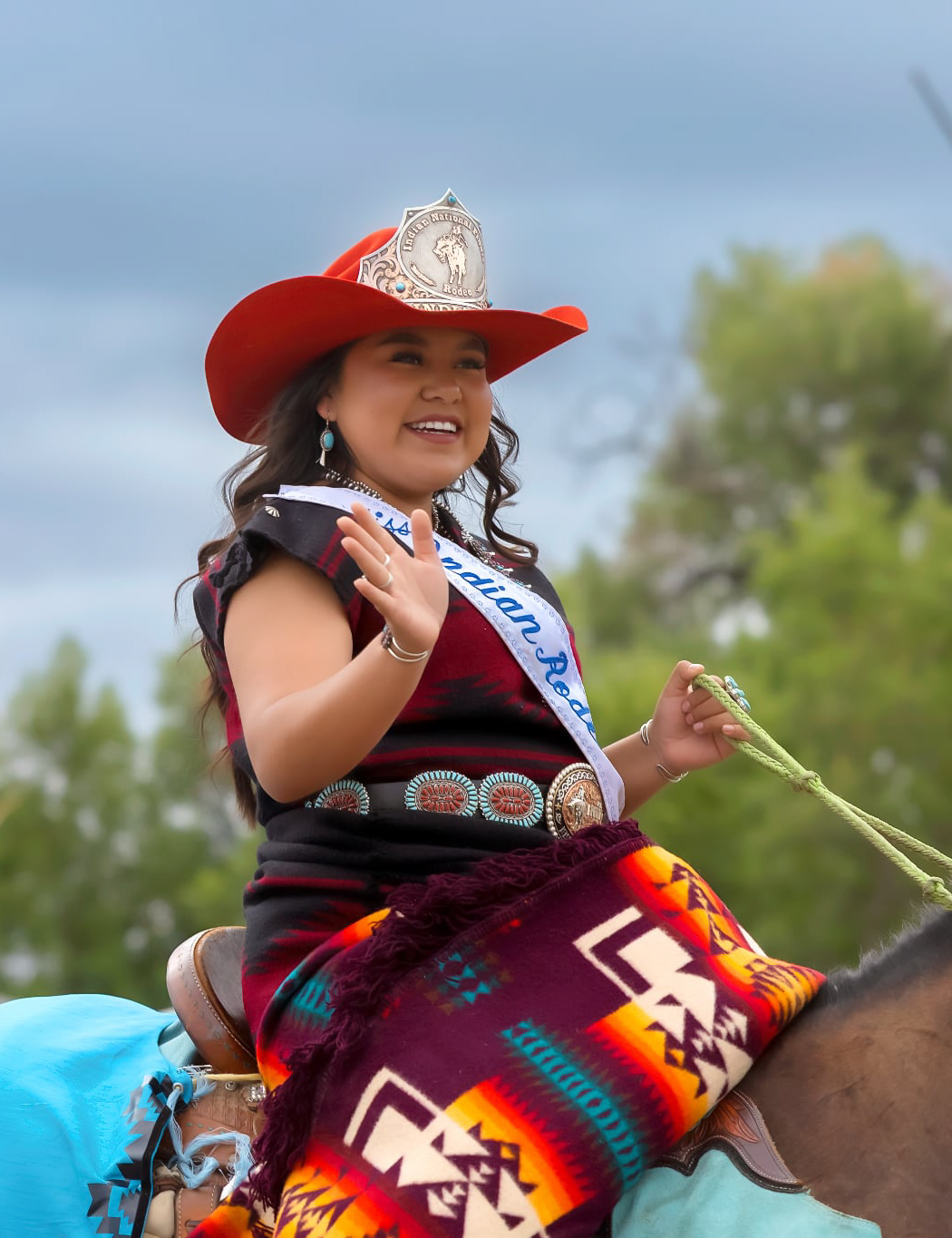 Native rodeo titleholder in a red cowboy hat, crown and sash at the Crow Fair Parade.
